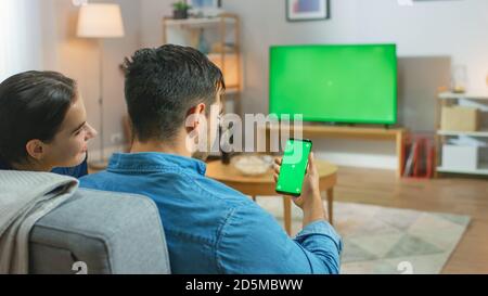 Una coppia felice che si siede a casa nel soggiorno guardando la TV Green Chroma con schermo a tasti, rilassandosi su un divano. Guy usa anche Green Mock-up schermo Foto Stock