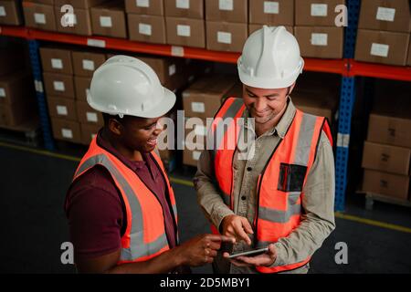 Due lavoratori maschi che scorrono sul tablet digitale discutono il processo di consegna dei pacchi confezionati in fabbrica. Foto Stock