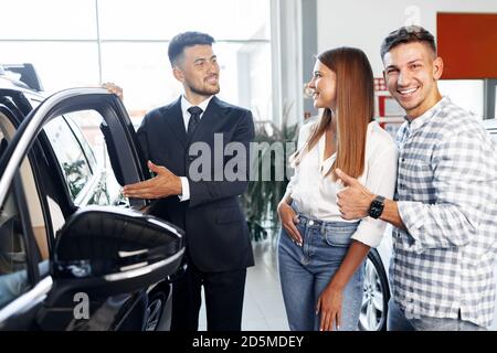 Giovane coppia che sceglie un'auto presso la concessionaria con il manager aiuto Foto Stock