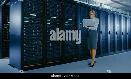 Nei data center: Il tecnico IT femminile si trova vicino all'armadietto rack ed esegue il programma di manutenzione sul notebook, controlla l'ottimale del server operativo Foto Stock