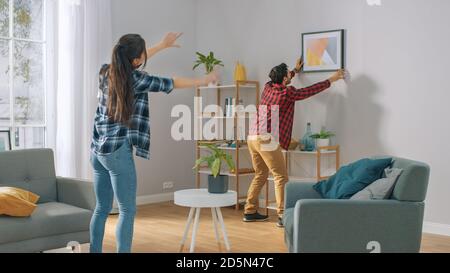 Happy Couple Hanging Picture on the Wall, Boyfriend si muove, la ragazza gli dice quando la cornice è dritto Hanging. Momento divertente in giovane coppia Foto Stock
