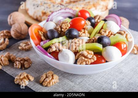 Insalata con pomodori ciliegini freschi, mozzarella, olive nere, kiwi, cipolla e noci su fondo di legno nero, pancake, primo piano. Foto Stock
