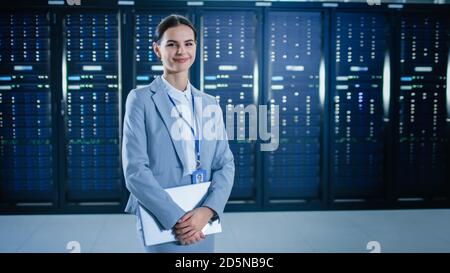 La Specialista IT femminile si sta levando in piedi nel data center accanto ai rack server, tenendo il laptop e sorridendo alla videocamera. Foto Stock
