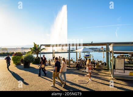 Le persone che passeggiano sul lago di Ginevra alla fine di una giornata di sole o si godono la terrazza di una gelateria con vista sulla fontana a getto d'acqua. Foto Stock