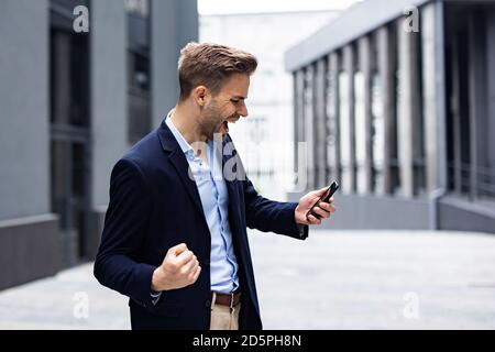 Eccitato giovane uomo che celebra la vittoria. Grande successo, grandi successi, emozioni positive. Bell'uomo d'affari con smartphone che ottiene buone notizie Foto Stock