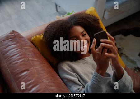 Vista verticale ad alto angolo di una donna africana allegra su cui giace divano utilizzando lo smartphone a casa Foto Stock