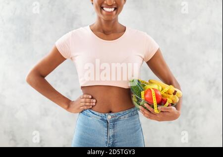 Vista in primo piano della giovane donna afroamericana che tiene frutta fresca e verdure con nastro di misurazione su sfondo grigio Foto Stock