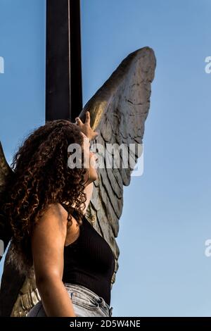 Colpo verticale di una bella donna che posa da un angelo statua con la mano su un'ala Foto Stock