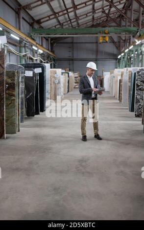 Operaio maschile a corpo pieno in un tablet di navigazione casco mentre si lavora in magazzino di marmo Foto Stock