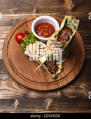 Burrito o tortilla serviti in ristorante o caffè. Pane piatto con ripieno di maiale o pollo e salsa di peperoncino rosso. Carne tritata avvolta con pane di lavasino su fondo di legno. Concetto di cucina tradizionale. Foto Stock