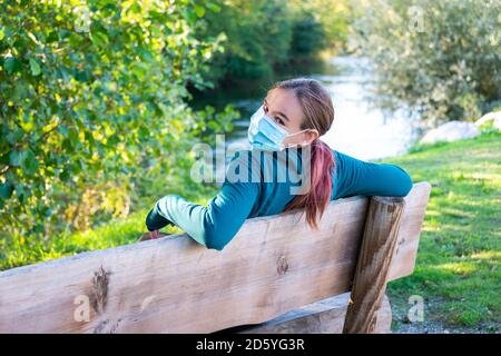 giovane donna seduta sulla panchina del parco che indossa una maschera accanto ad un fiume da solo; Foto Stock