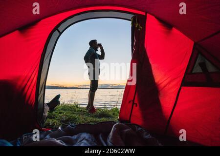 Uomo in campeggio in Estonia, in piedi di fronte alla tenda, guardando il tramonto Foto Stock