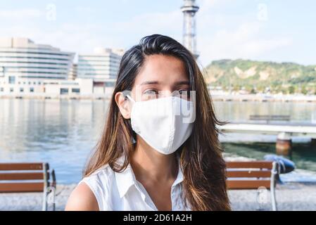 ritratto di una giovane donna indiana vestendo casual, indossando una maschera igienica bianca e guardando la macchina fotografica vicino al porto Foto Stock