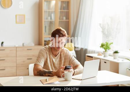 Uomo maturo seduto al tavolo di fronte al computer portatile e utilizzando il telefono cellulare in camera Foto Stock