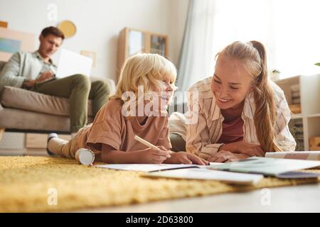 Madre felice che si stesa sul pavimento insieme al figlio e si disegnano con il padre seduto sul divano in sfondo Foto Stock