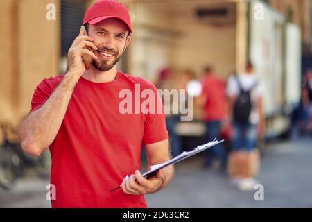 Giovane e felice courier maschio caucasico con tablet che parla al telefono mentre si trova in deposito cortile. Concetto di consegna degli alimenti Foto Stock