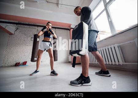 Una giovane ragazza di kickboxer forte calcia il suo partner scintillante con una zampa di pugilato in una sessione di allenamento personale Foto Stock