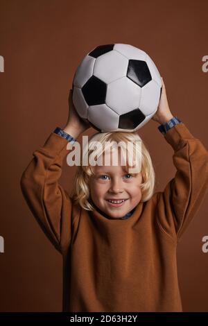 Ritratto di bambino carino con i capelli biondi che tengono il calcio sopra la sua testa e guardando la macchina fotografica contro lo sfondo marrone Foto Stock
