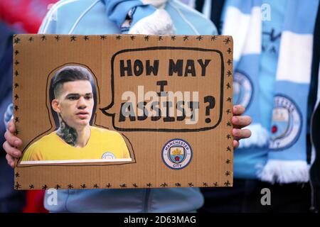 Un fan di Manchester City tiene un banner negli stand con il portiere di Manchester City, Ederson, che legge "come posso assistere?" Foto Stock