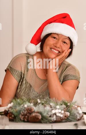 Donna asiatica felice osservante matura con un cappello di Santa seduta Ad un tavolo con le decorazioni di Natale Foto Stock
