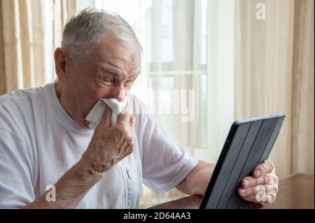 l'uomo anziano con allergie e tosse e starnuti freddi si trasforma in un tovagliolo. Un anziano malato lavora su un computer mentre si trova a casa durante l'epidemia Foto Stock