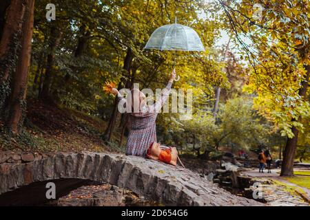 Felice giovane donna seduta sul margine da stagno nel parco autunnale alzando ombrello trasparente durante la pioggia. Passeggiata nella stagione autunnale Foto Stock