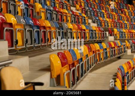 File di sedili ripiegati, verdi, blu, gialli, verdi e arancioni in plastica in uno stadio interno molto grande e vuoto Foto Stock