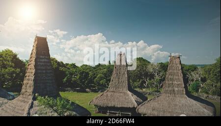 Luce del sole sul tetto di paglia alta della casa tradizionale nel villaggio di Kodi, Indonesia. Vista aerea del primitivo insediamento indonesiano addmist verde foresta tropicale. Splendida vacanza estiva in Asia unico punto di riferimento Foto Stock