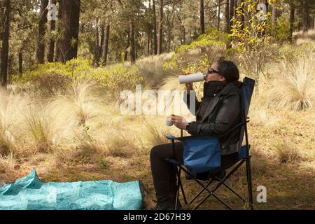 Donna seduta sulla sedia che ha un picnic nella foresta godendo il suo drink preferito mentre nella foresta circondata da alberi di pino e prateria gialla in Foto Stock