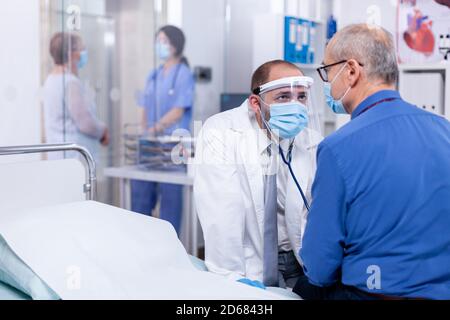 Esame del battito cardiaco del paziente con stetoscopio nella sala dell'ospedale e uso della maschera facciale come precauzione di sicurezza contro il coronavirus. Visita medica per infezioni, malattie e diagnosi. Foto Stock