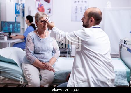 Personale medico che utilizza un termometro a infrarossi per prendere la temperatura corporea della paziente anziana. Consulenza medica per infezioni e malattie durante pandemia globale, influenza, strumento, malattia. Foto Stock