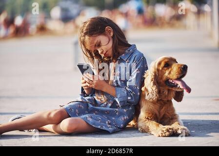 Siede sul pavimento. Con lo smartphone nelle mani. Carina bambina hanno una passeggiata con il suo cane all'aperto in giornata di sole Foto Stock