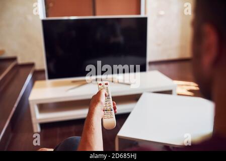 La vista posteriore di MAN tiene il telecomando in mano e. Accendere il televisore Foto Stock