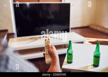 Con birra sul tavolo. La vista posteriore di MAN tiene il telecomando in mano e accende la TV Foto Stock