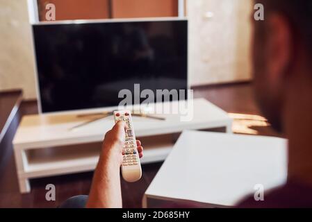 La vista posteriore di MAN tiene il telecomando in mano e. Accendere il televisore Foto Stock