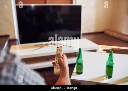Con birra sul tavolo. La vista posteriore di MAN tiene il telecomando in mano e accende la TV Foto Stock