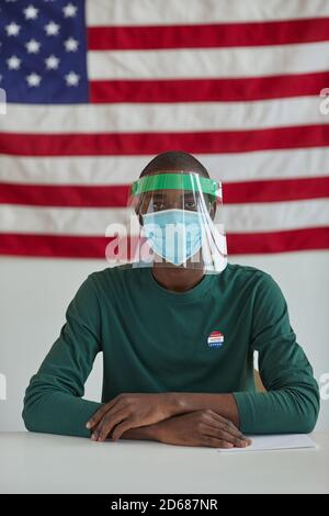 Ritratto dell'uomo africano in maschera protettiva seduto al Tavolo durante il giorno delle elezioni con bandiera americana in background Foto Stock