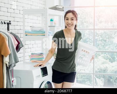 Bella giovane donna asiatica casalinga in piedi e tenendo bianco vuoto cestino di stoffa con sorriso vicino lavatrice in lavanderia vicino alla finestra con Foto Stock