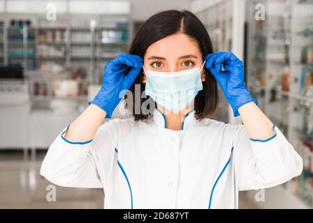 chiudi ritratto femmina farmacista in farmacia mettendo sul viso a maschera protettiva che si sta lavorando per aiutare le persone Foto Stock