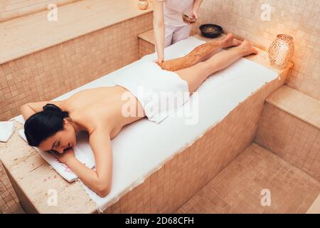 Spa salon operaio che applica scrub sulle gambe di un cliente salone di bellezza. Esfoliare la pelle più vecchia dei piedi con uno scrub corpo. Procedura di spellatura della brunetta Foto Stock