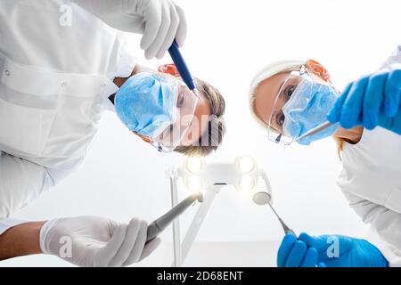 Dentista e assistente che indossa occhiali di protezione e maschera medica che regge l'attrezzatura dentale. Trattamento denti, vista dal basso. Lavoro di squadra di odontoiatria Foto Stock