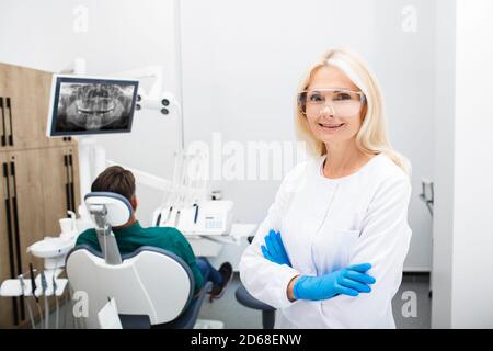 Ritratto di una donna odontoiatra sorridente, in occhiali protettivi a mani incrociate, ambulatorio di odontoiatria di fondo con un paziente Foto Stock