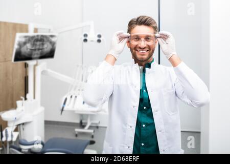 Ritratto di un bel dentista maschile sorridente che indossa occhiali protettivi in una moderna clinica dentale. Odontoiatria moderna Foto Stock