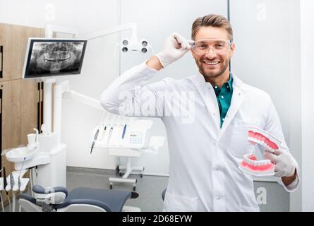 Ritratto di un bel dentista che sorride e guarda la macchina fotografica, tenendo un modello a denti. Trattamento indolore delle carie in un moderno studio dentistico Foto Stock