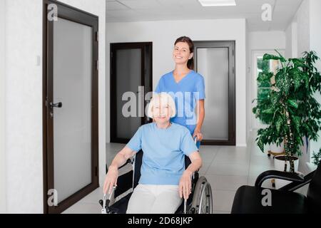 Infermiere amichevole che si prende cura del paziente anziano in un sedia a rotelle nel corridoio dell'ospedale Foto Stock