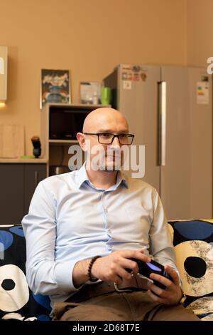 Uomo caucasico mid adult che gioca a videogiochi a casa. Uomo felice seduto sul divano con joystick o console di gioco in mani Foto Stock