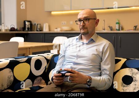 l'uomo calvo di mezza età con gli occhiali sta giocando i video giochi con il joystick a casa sul divano. L'uomo d'affari allevia lo stress dopo il lavoro Foto Stock