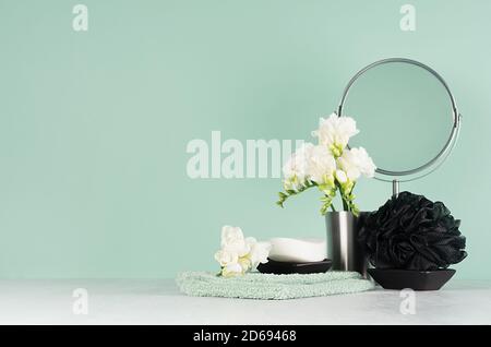 Elegante arredamento bagno con accessori neri per il bagno con fiori bianchi primaverili, specchio, sapone, asciugamano di cotone su tavolo di legno bianco, parete di menthe alla menta. Foto Stock