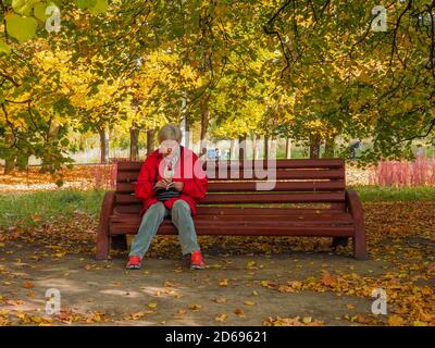 Mosca. Russia. Il 3 ottobre 2020, una donna anziana dai capelli grigi in una giacca rossa si siede su una panchina del parco sotto un albero e legge un libro. Soleggiato giorno d'autunno Foto Stock
