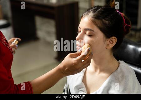 Una bella giovane ragazza indiana ottiene il trucco fatto ad un salone, l'artista del trucco che applica la spugna sulla faccia del modello, trucco e concetto di salone di bellezza. Foto Stock